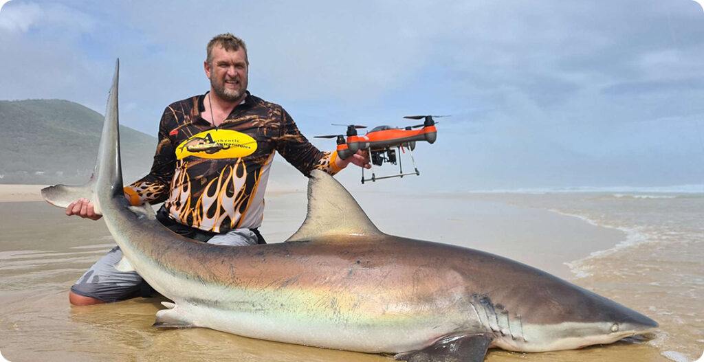 Land-based shark fishing with fisherman kneeling next to a large shark and holding his SwellPro drone