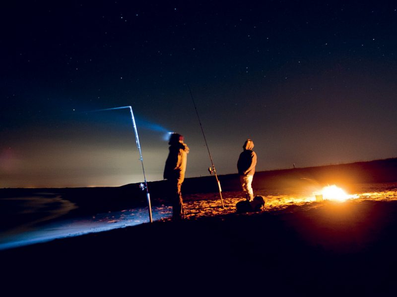 surf fishing safety - nighttime surf fishing two anglers next to their campfire watching their rods