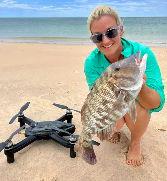 Lady angler kneeling on the beach next to her Cuta Copter Trident Boss+ waterproof fishing drone while holding a nice catch.