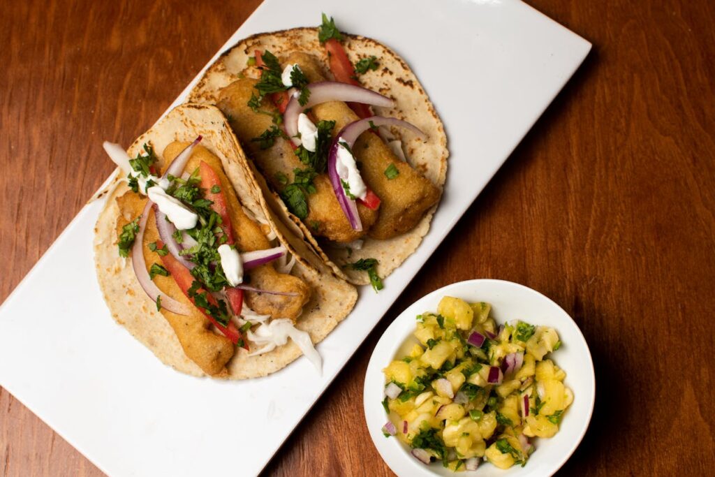 Two Fish tacos on a rectangular plate with a side of pineapple salsa in a small bowl.
